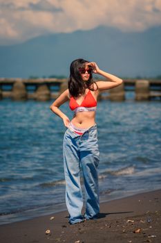 Stylish woman in red bra and jeans poses on a scenic beach with mountains in the background.