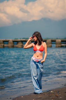 Fashionable woman poses at a picturesque beach, wearing a red bikini and sunglasses, with mountains in the backdrop.