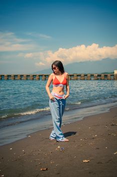 Woman posing in a red bikini top and jeans on a sandy beach under a clear blue sky.
