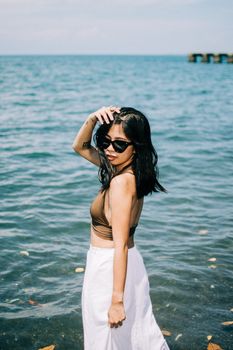 Fashion-forward woman in sunglasses stands by the sea in summer attire.