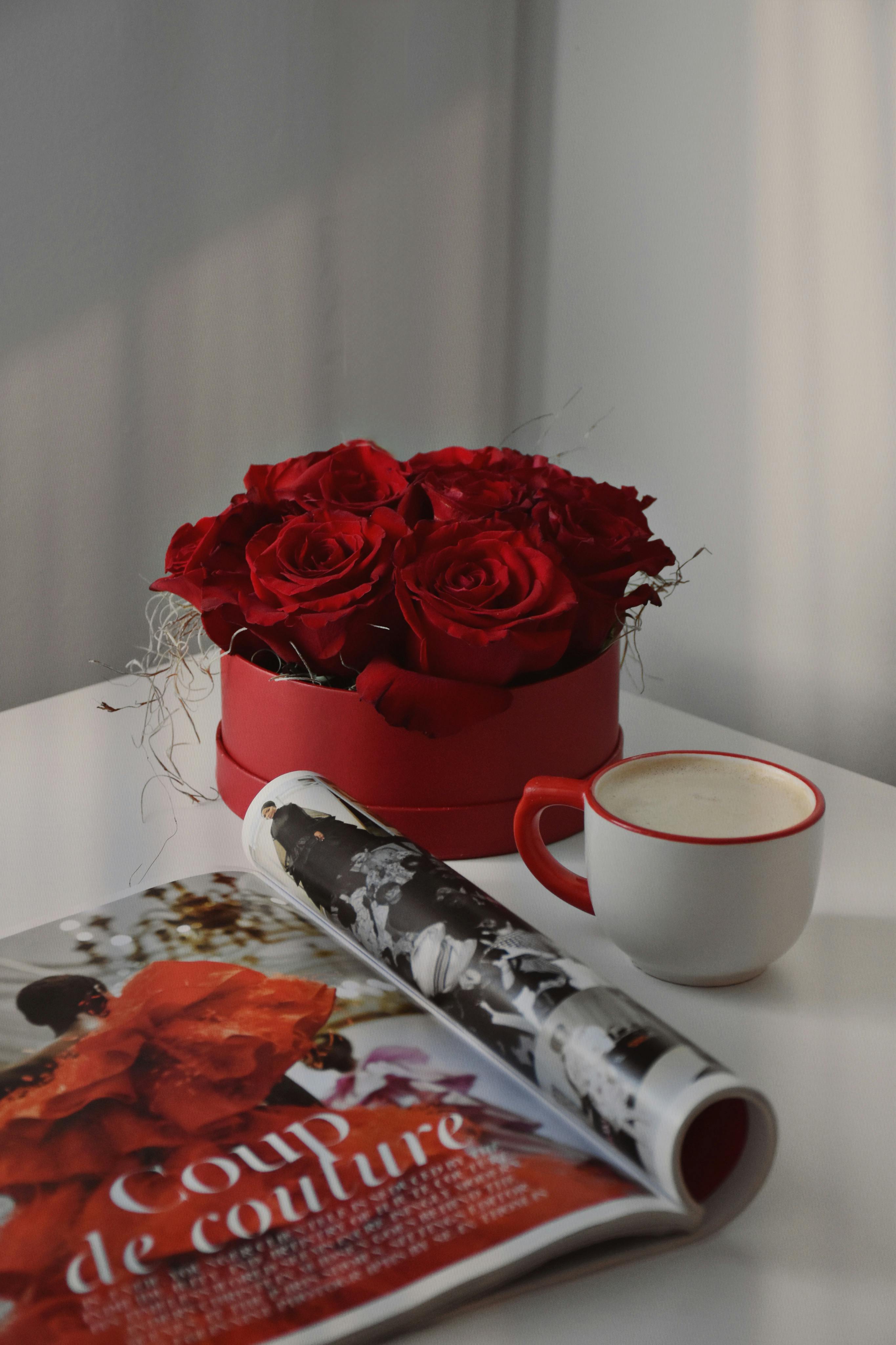 A stylish arrangement of red roses beside a cappuccino and a fashion magazine on a sunlit table.