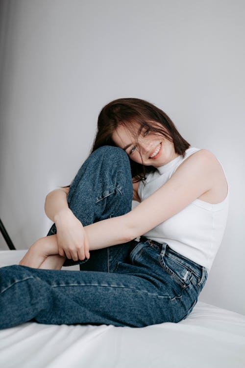 Young Woman Sitting on a Bed in Blue Jeans and White High Collar Top