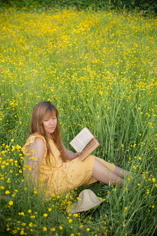 A Woman in a Yellow Dress Reading a Book on a Meadow with Yellow Flowers