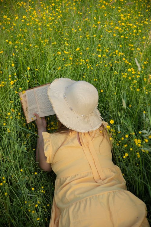 Free A woman in a yellow dress lies in a sunny meadow surrounded by yellow flowers, reading a book. Stock Photo