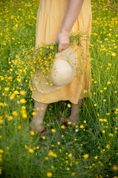 A woman in a yellow dress holding a hat and flowers in a vibrant summer meadow.