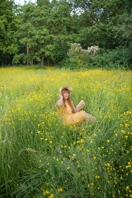 A Woman in a Yellow Dress Reading a Book on a Meadow with Yellow Flowers