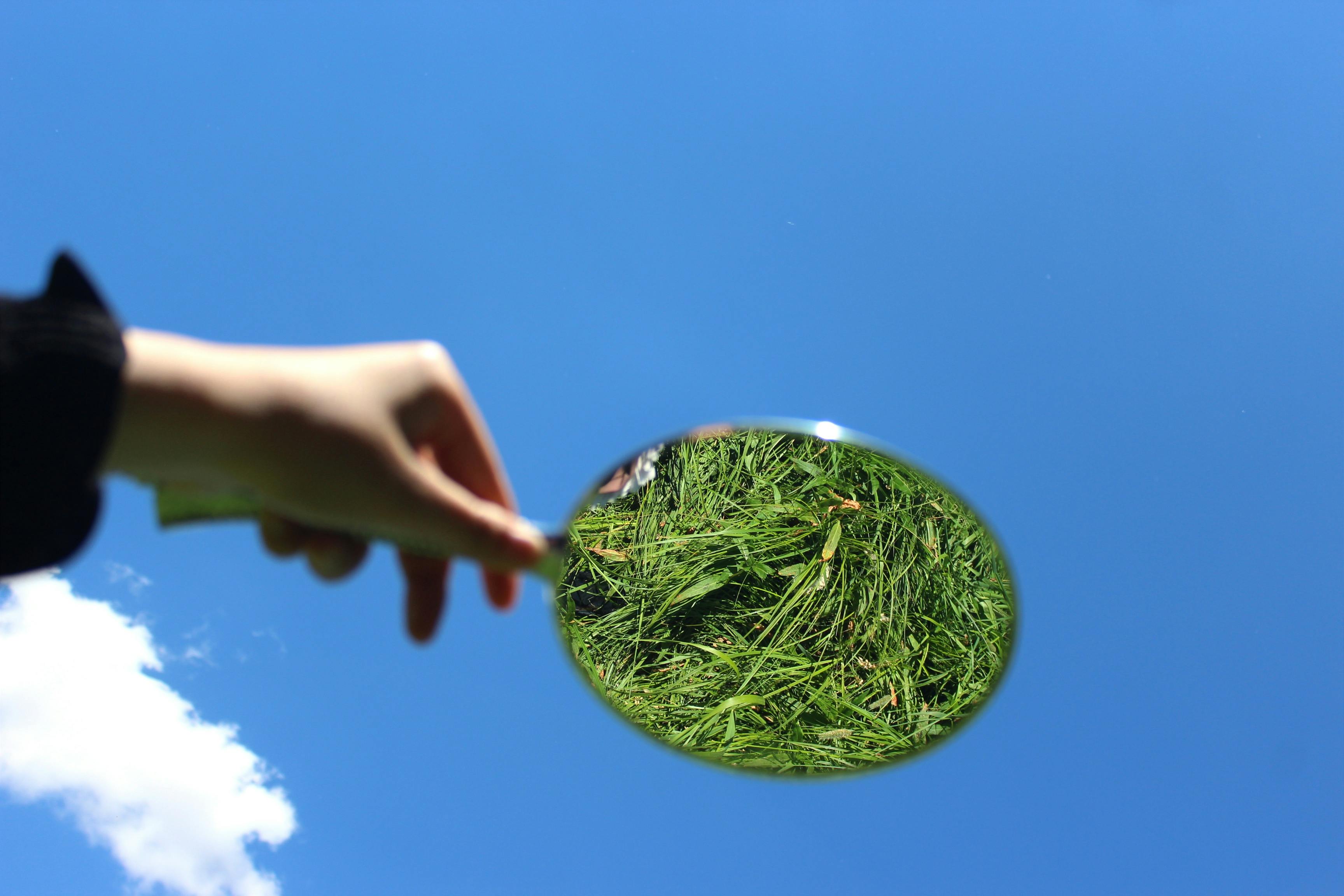 A hand holds a mirror reflecting grass, set against a vibrant blue sky backdrop.