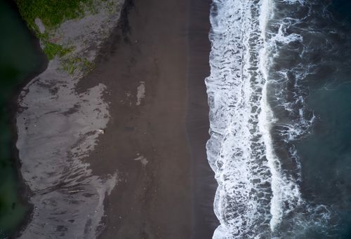 Free Drone shot capturing the serene beauty of waves crashing on a sandy beach with lush greenery. Stock Photo