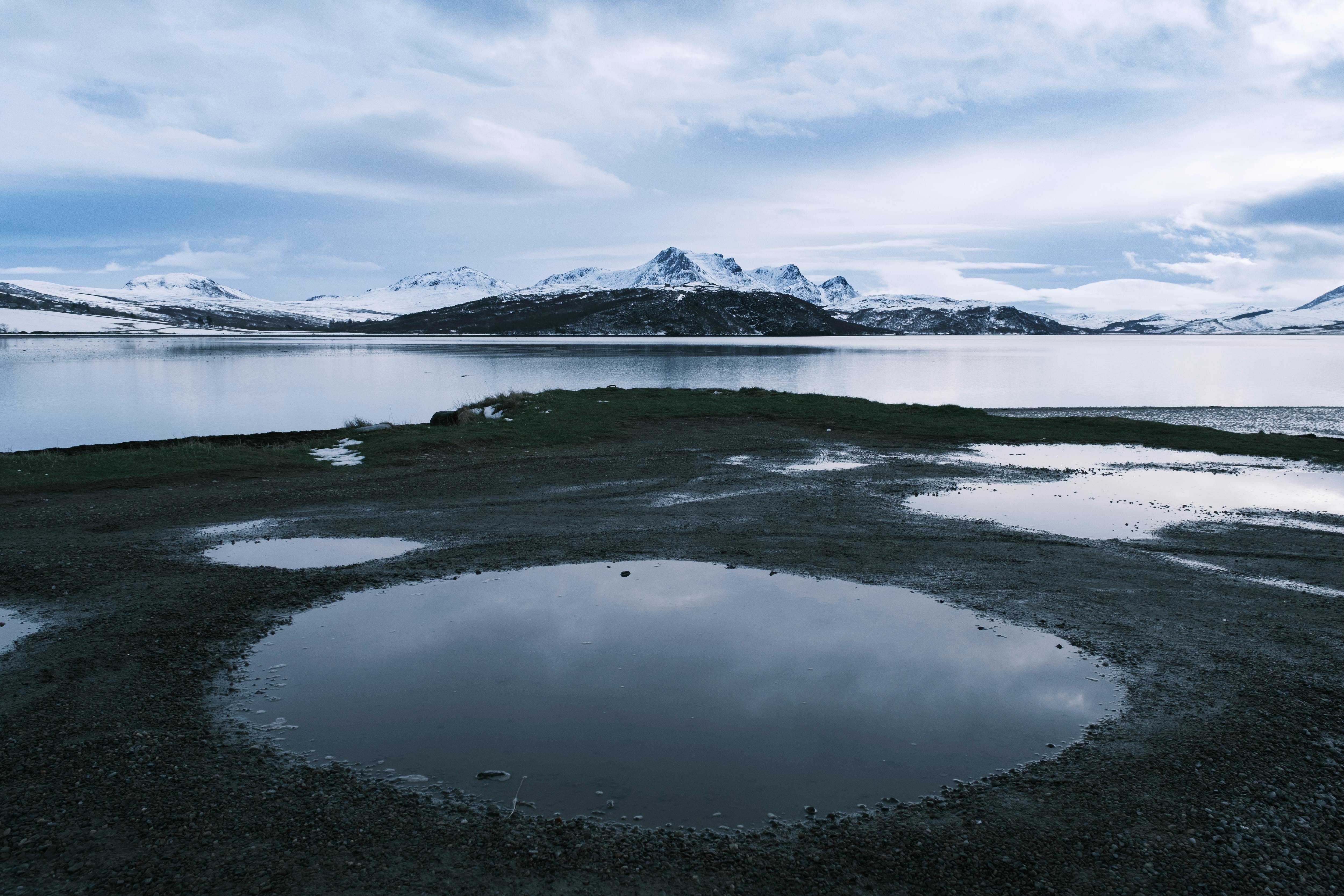 A serene winter landscape with mountains reflected in a puddle by a lakeside.