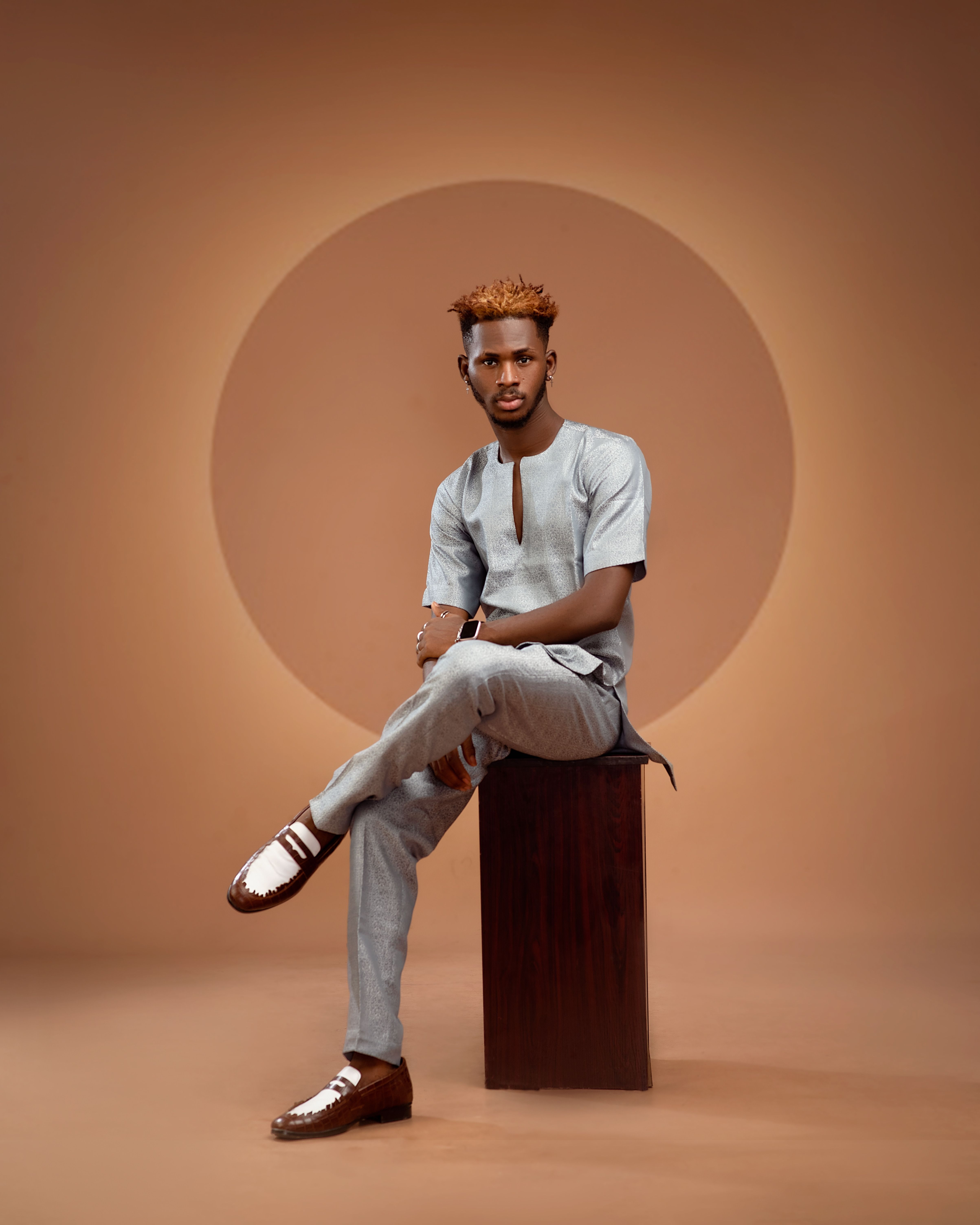 Fashionable man with dyed hair and stylish outfit sitting on a stool against a brown backdrop.