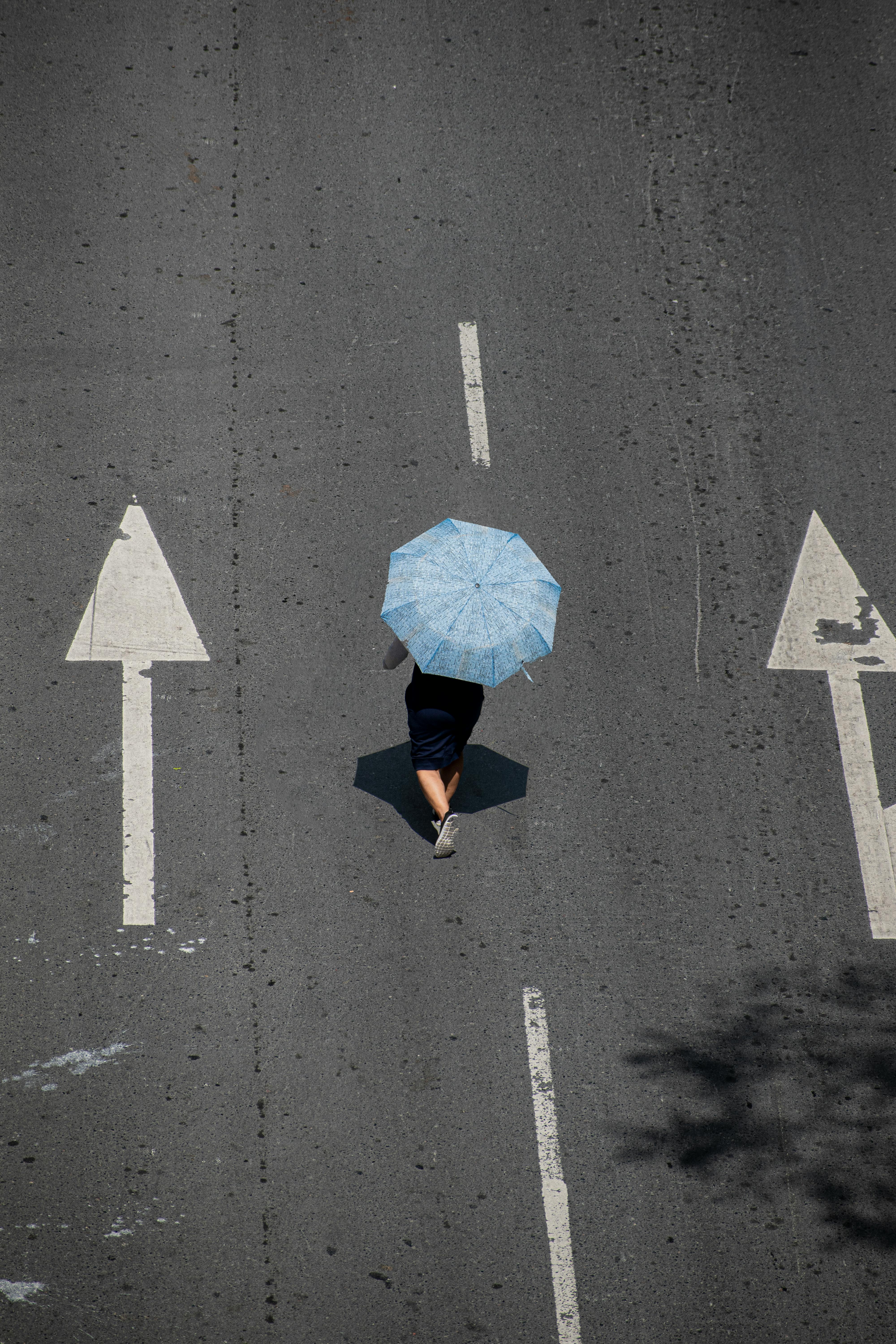 Aerial shot of a person with a blue umbrella walking between road markings in an urban setting.