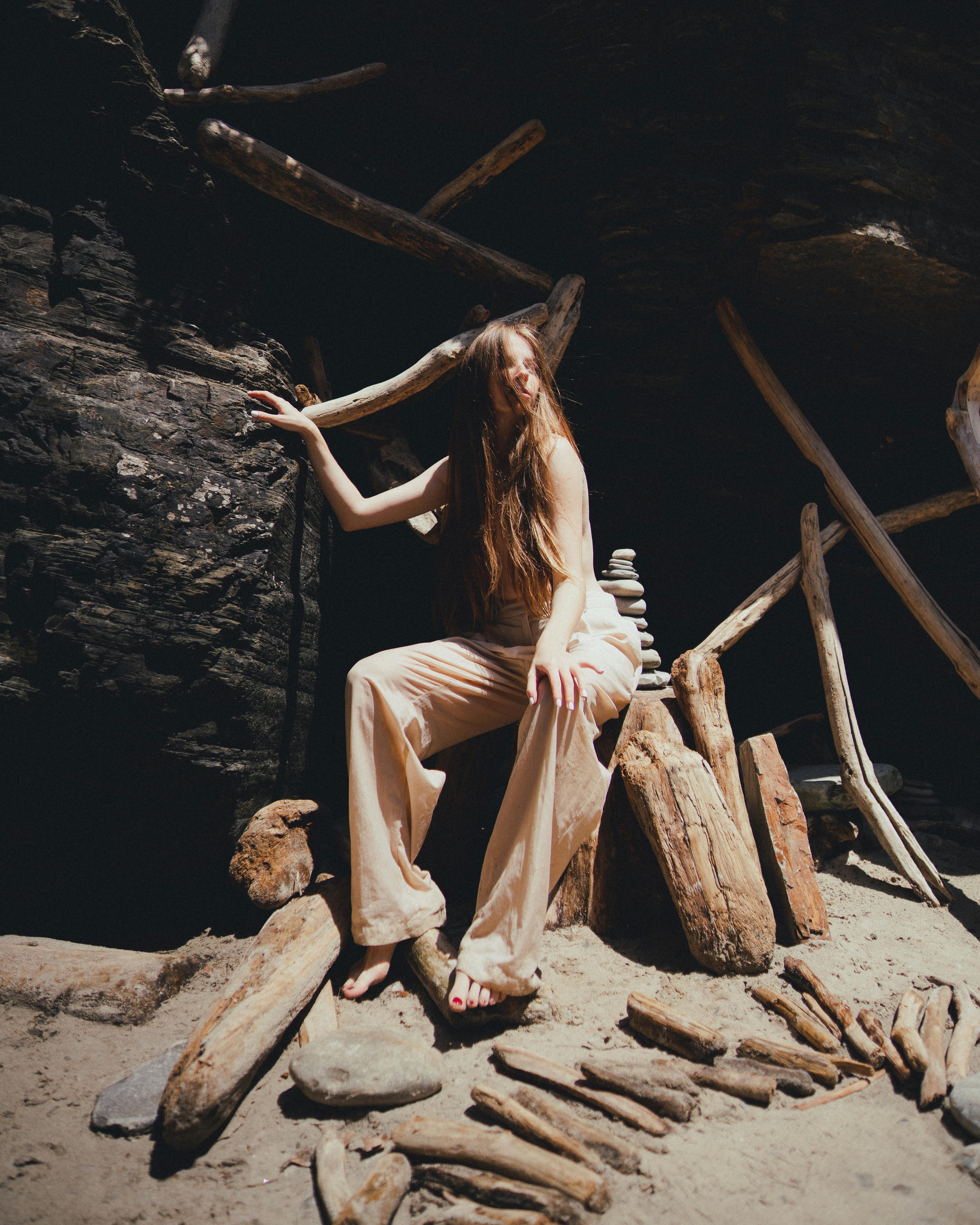 A woman in stylish attire poses in a rustic, artistic outdoor setting of logs and rocks.