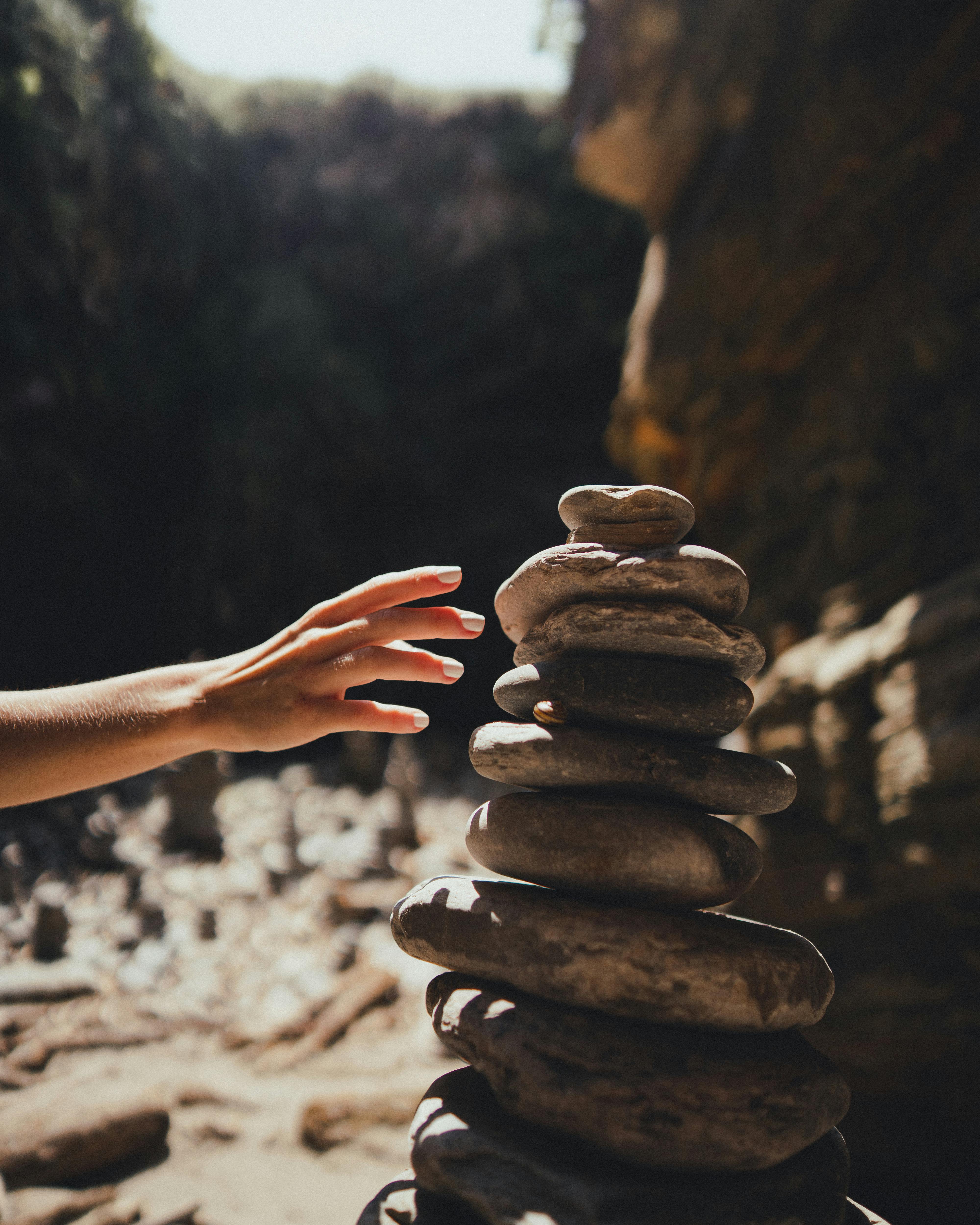 A hand reaches towards a carefully balanced stack of stones in a sunlit cave environment.