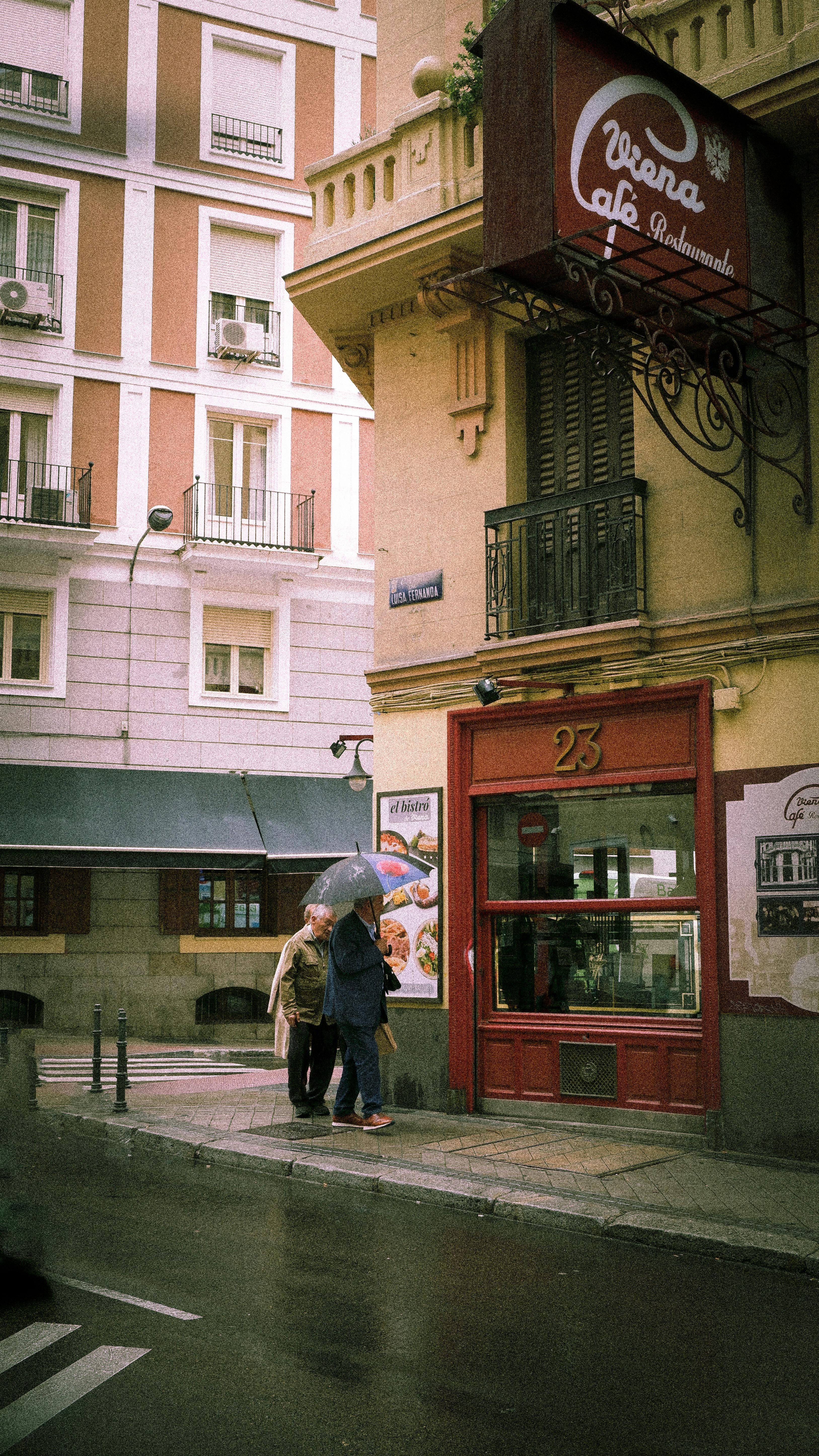 Free Charming city street scene with people under umbrellas by a cafe on a rainy day. Stock Photo