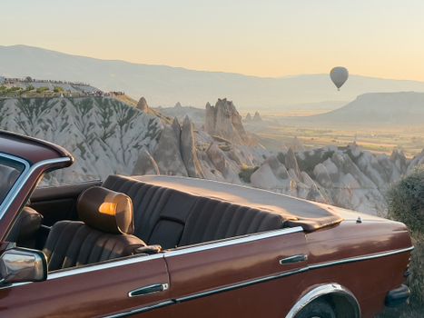 A vintage cabriolet parked at sunrise in Cappadocia with rock formations and a hot air balloon in the background.