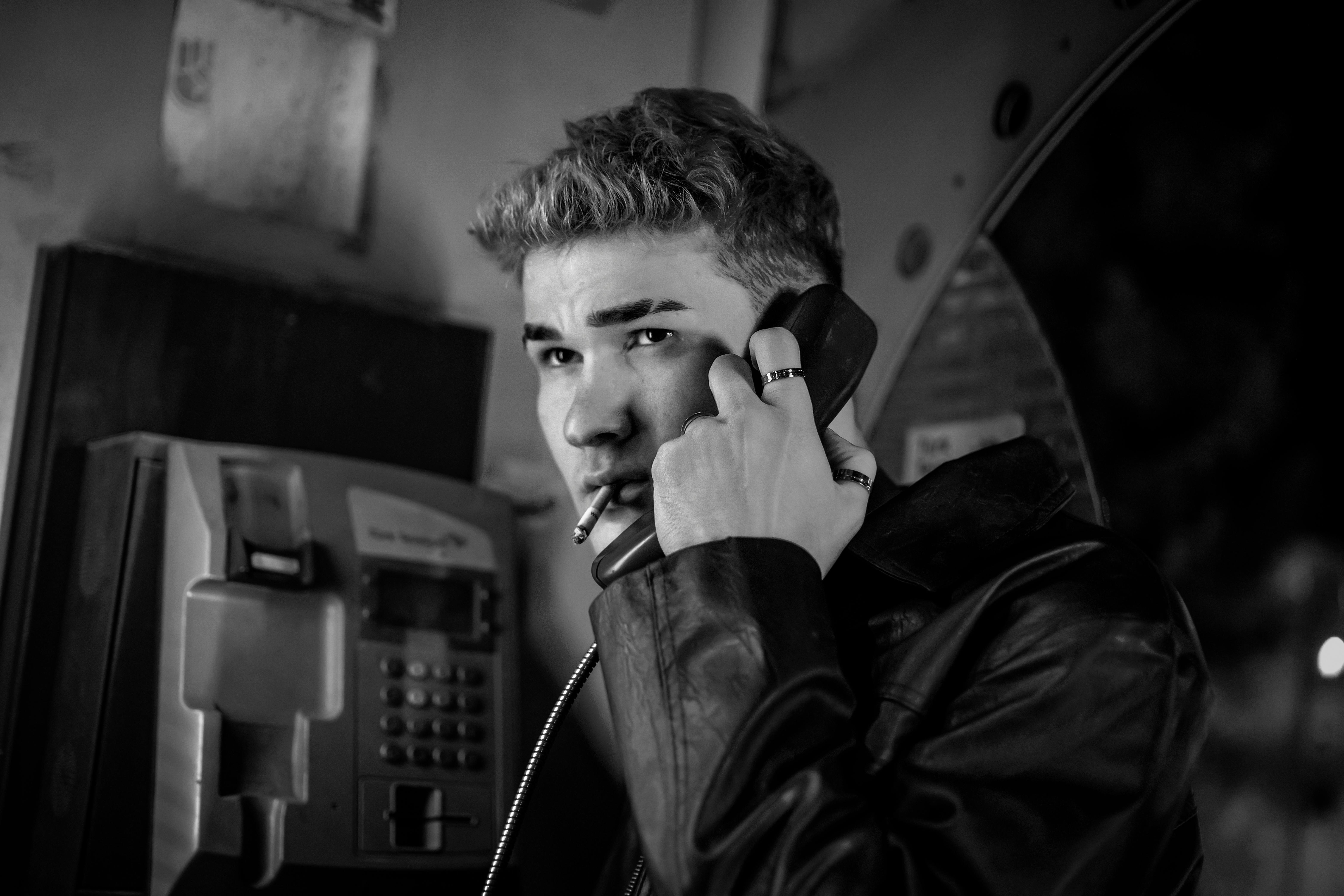 Black and white photo of a young man using a payphone, conveying a vintage feel and moody atmosphere.