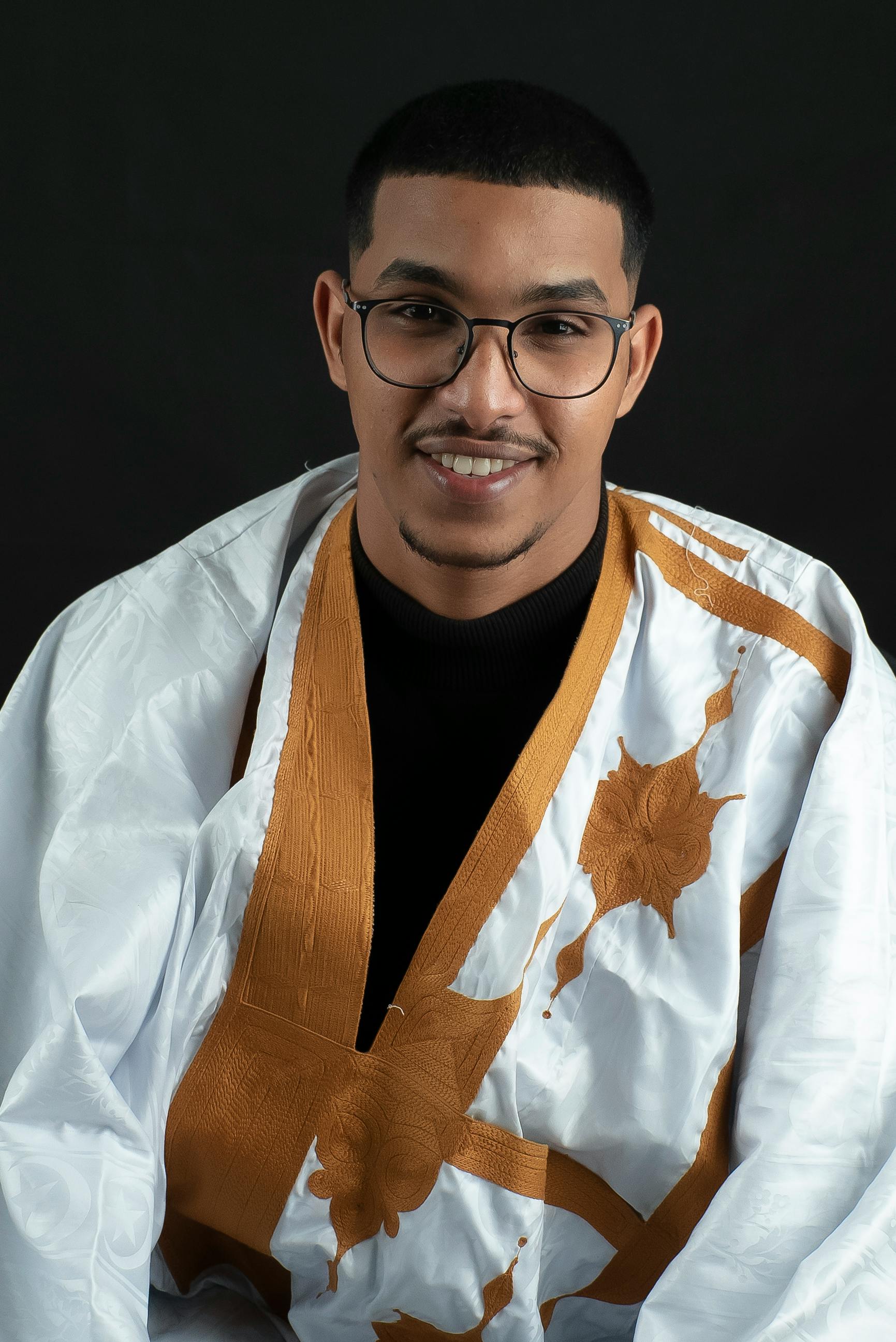 A smiling man wearing eyeglasses and traditional attire with a dark background.