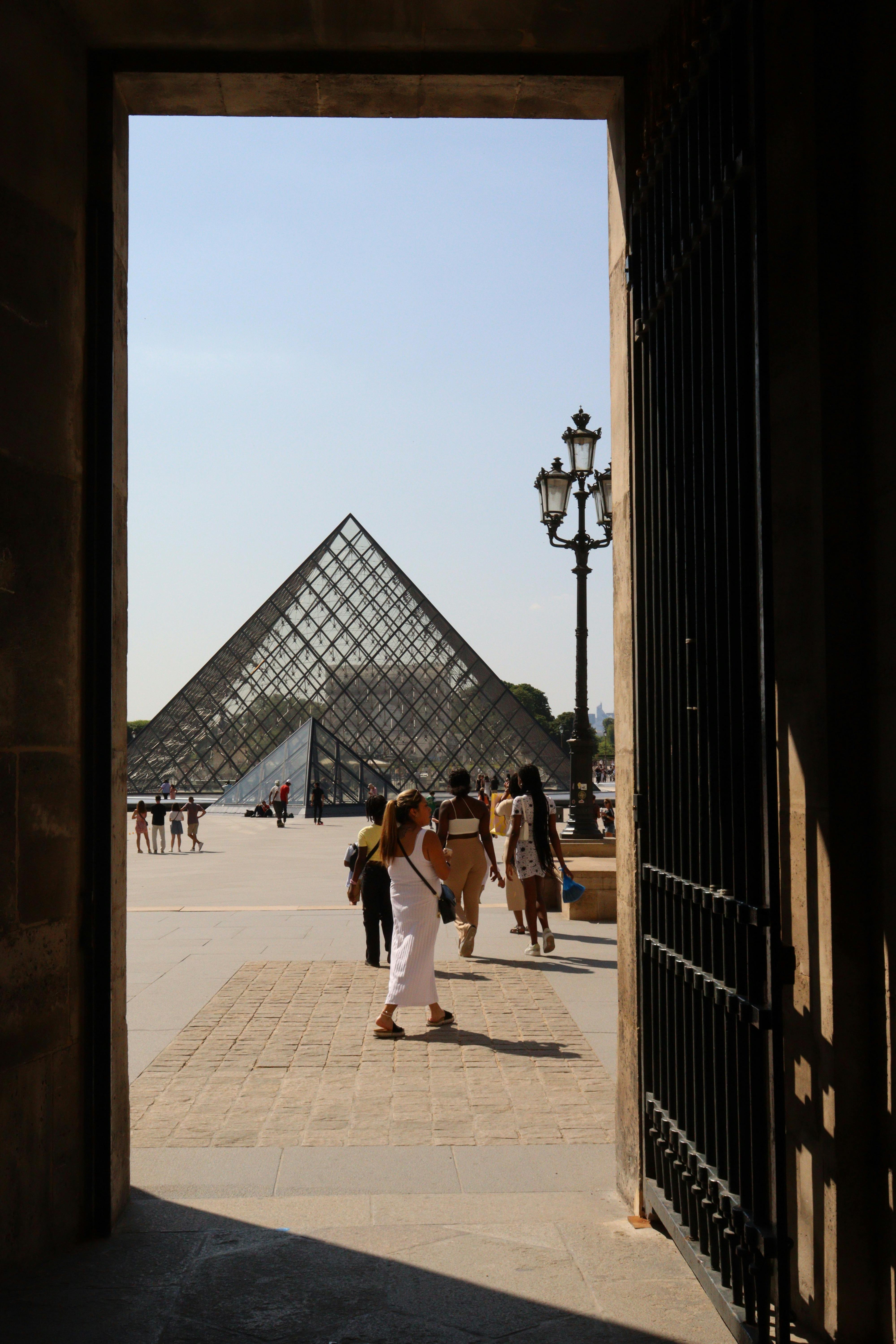 Visitors entering the Louvre courtyard, framed by an archway, with the iconic glass pyramid in view.