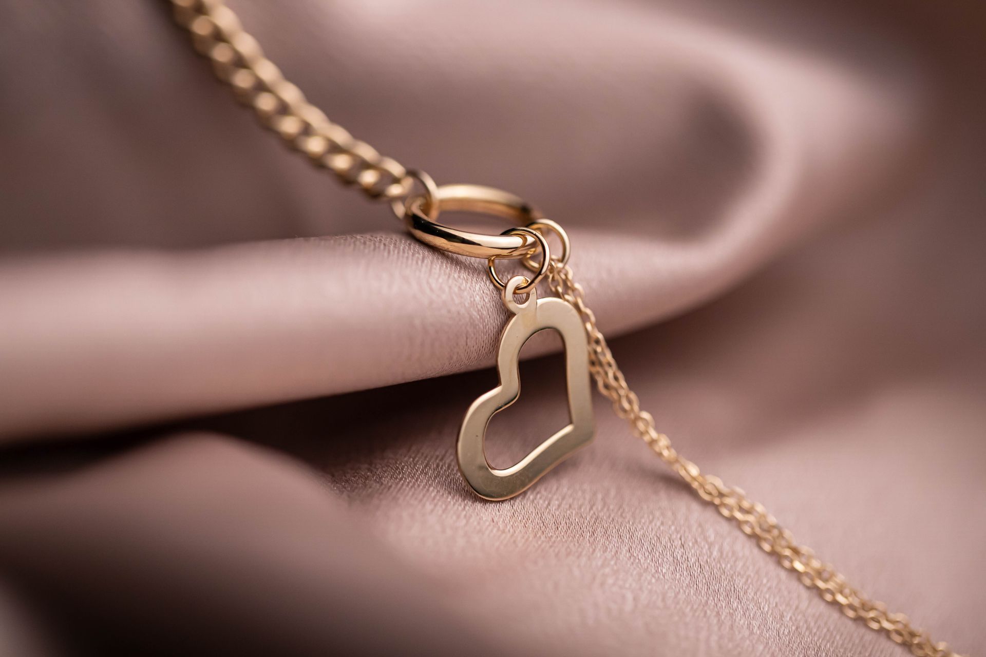 Close-up of a gold heart pendant necklace elegantly draped over soft, pink fabric with a delicate link chain.