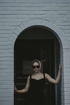 Stylish woman in sunglasses and headscarf posing under a brick archway in an urban setting.