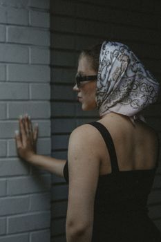 A stylish woman with a headscarf and sunglasses touches a brick wall, captured in a dramatic fashion shot.