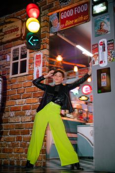 Fashionable woman in neon pants strikes a pose in a retro-themed diner setting at night.