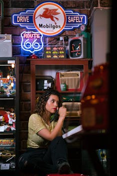 Woman sitting in a retro cafe with vintage decor, featuring neon signs and nostalgic items.