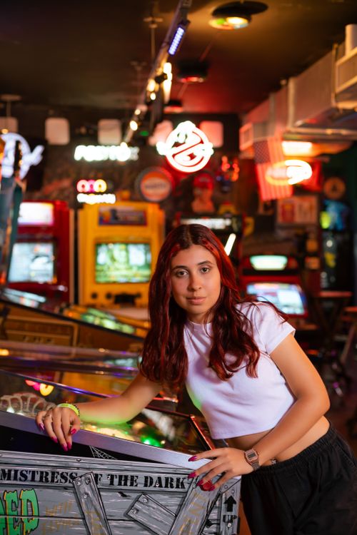 Free A young woman with long hair poses in a vibrant retro arcade setting filled with colorful lights. Stock Photo