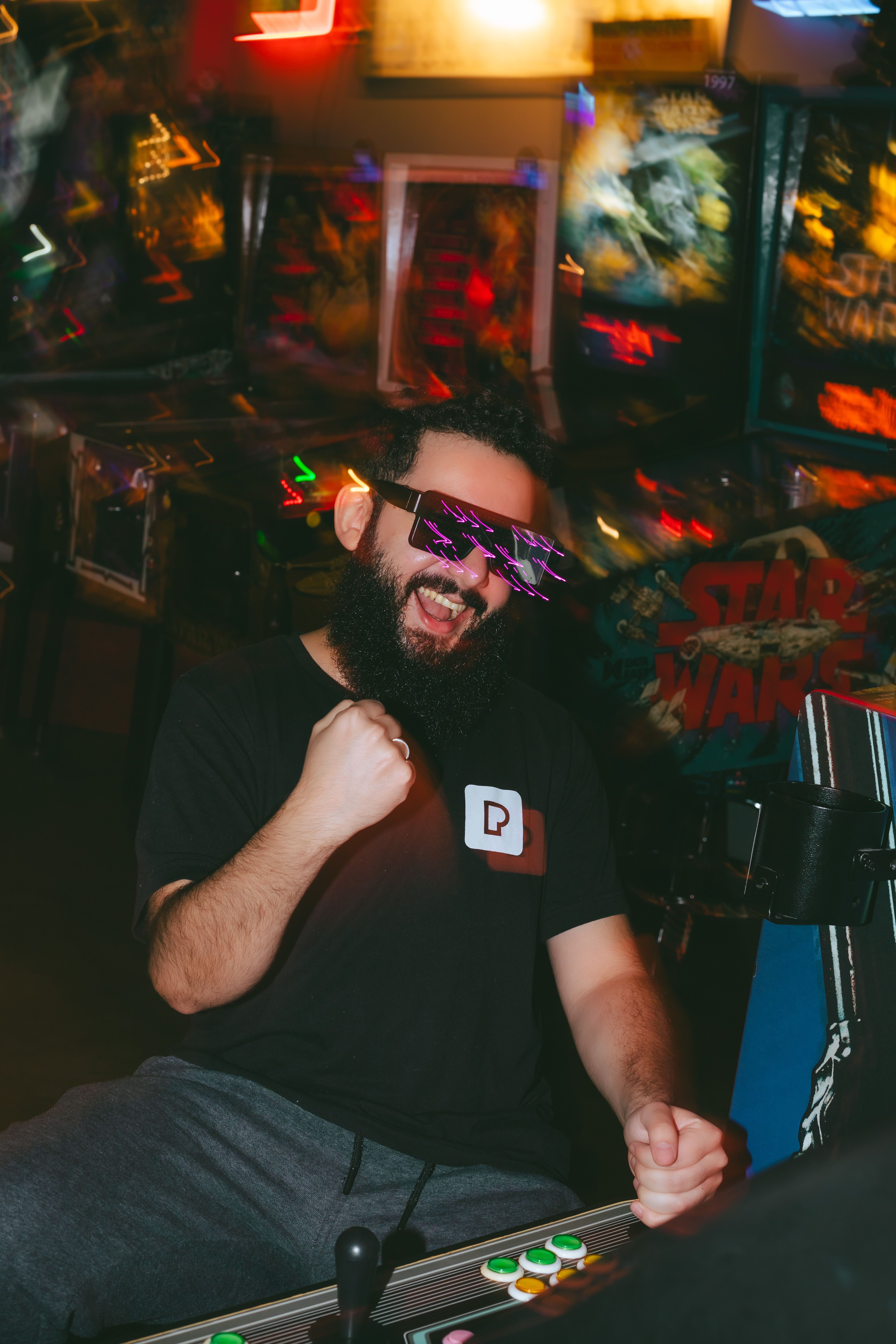 Bearded man wearing sunglasses celebrates in a lively arcade setting with vibrant lights.