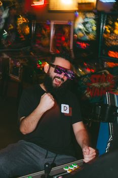 Bearded man wearing sunglasses celebrates in a lively arcade setting with vibrant lights.
