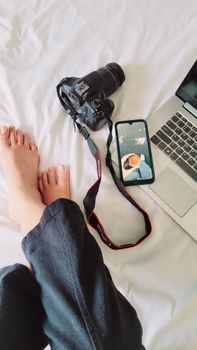 Woman relaxing with camera, laptop, and smartphone on white bed. Ideal for modern lifestyle themes.