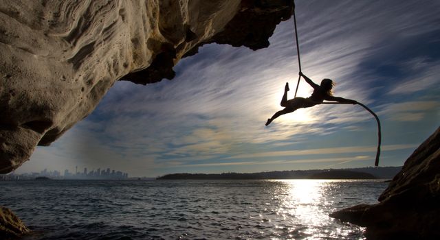 Silhouette of a woman performing acrobatics on a rope hanging from a cliff by the sea with city skyline in the background.