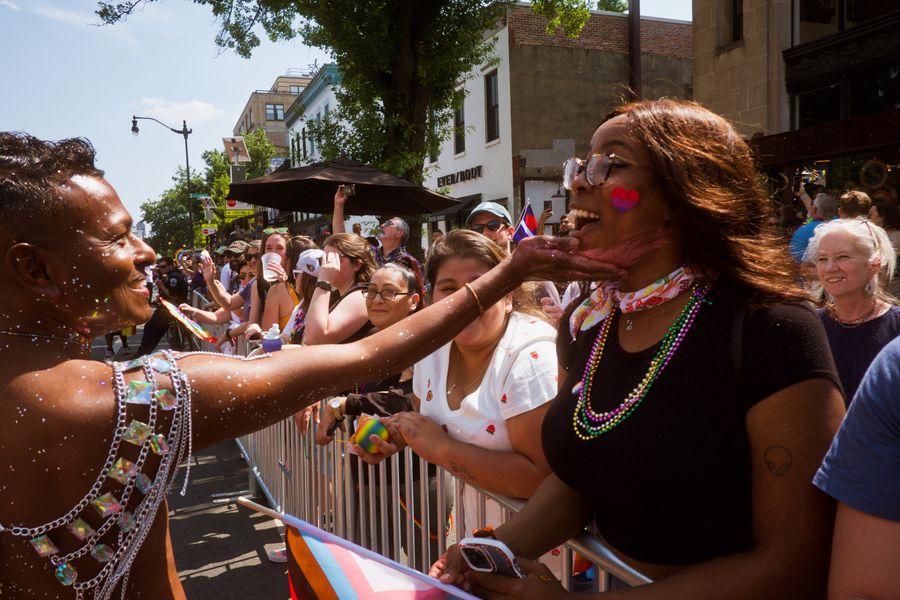 A crowd at a pride parade, with a performer in glittery attire touching a smiling attendee's face near a barricade, rainbow and progress-pride flags visible; location not identifiable as Piedmont Park.