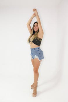 Portrait of a young woman posing confidently in a studio setting wearing denim shorts and a crop top on a white background.
