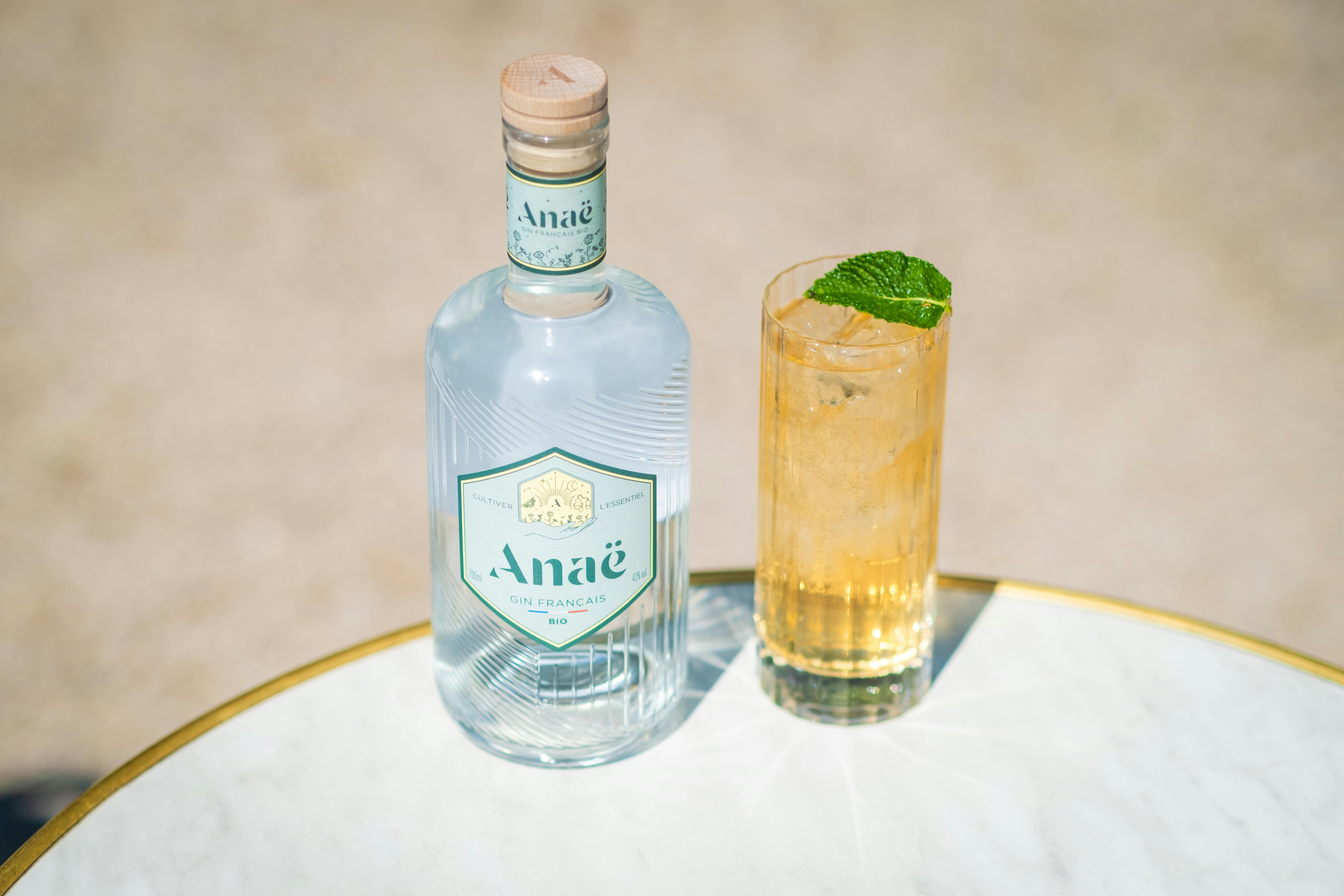 A crisp gin cocktail with mint garnish sits next to a stylish bottle on a marble table in bright daylight.