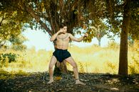 A topless man with tattoos striking a boxing pose in a sunny, rural outdoor area with trees.