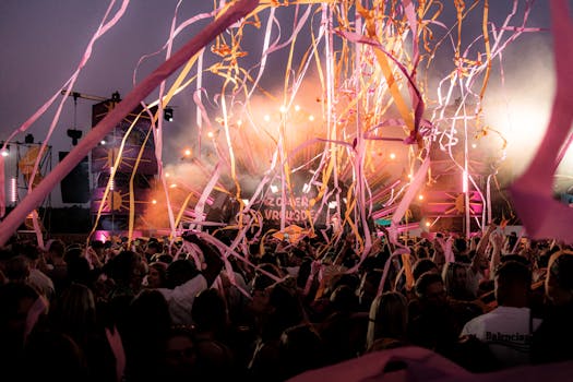 Crowd enjoying a vibrant festival with colorful ribbons and stage lights under a twilight sky.