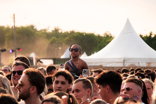 Excited crowd at an outdoor music festival with tents and glowing sunset ambiance.