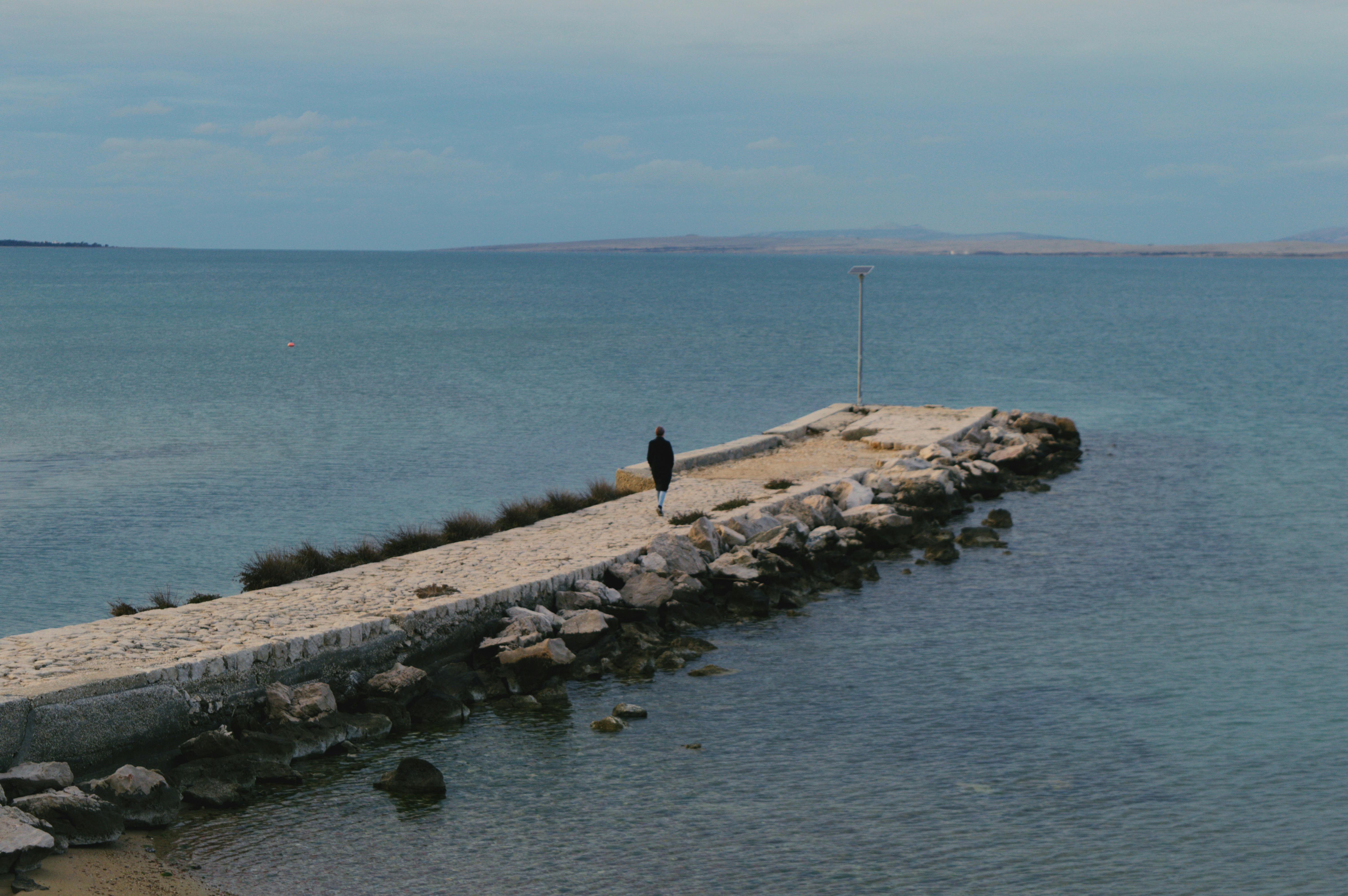 A person walks alone on a stone pier against a serene seascape, embodying solitude and calm.