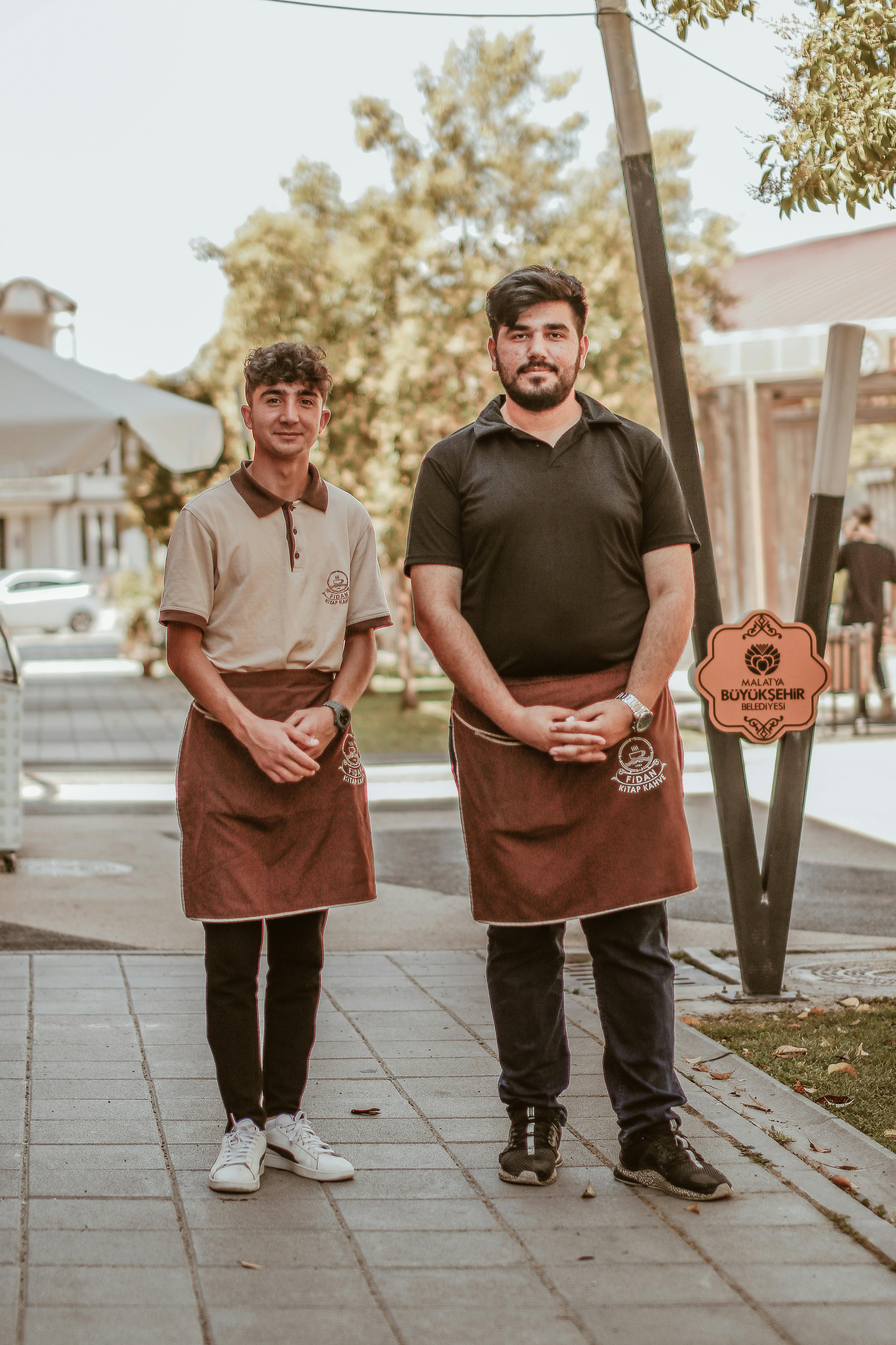 Two waiters posing on a city sidewalk, wearing aprons and smiling.