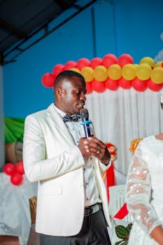 A man in a white suit speaking into a microphone during a festive indoor ceremony with balloon decorations.