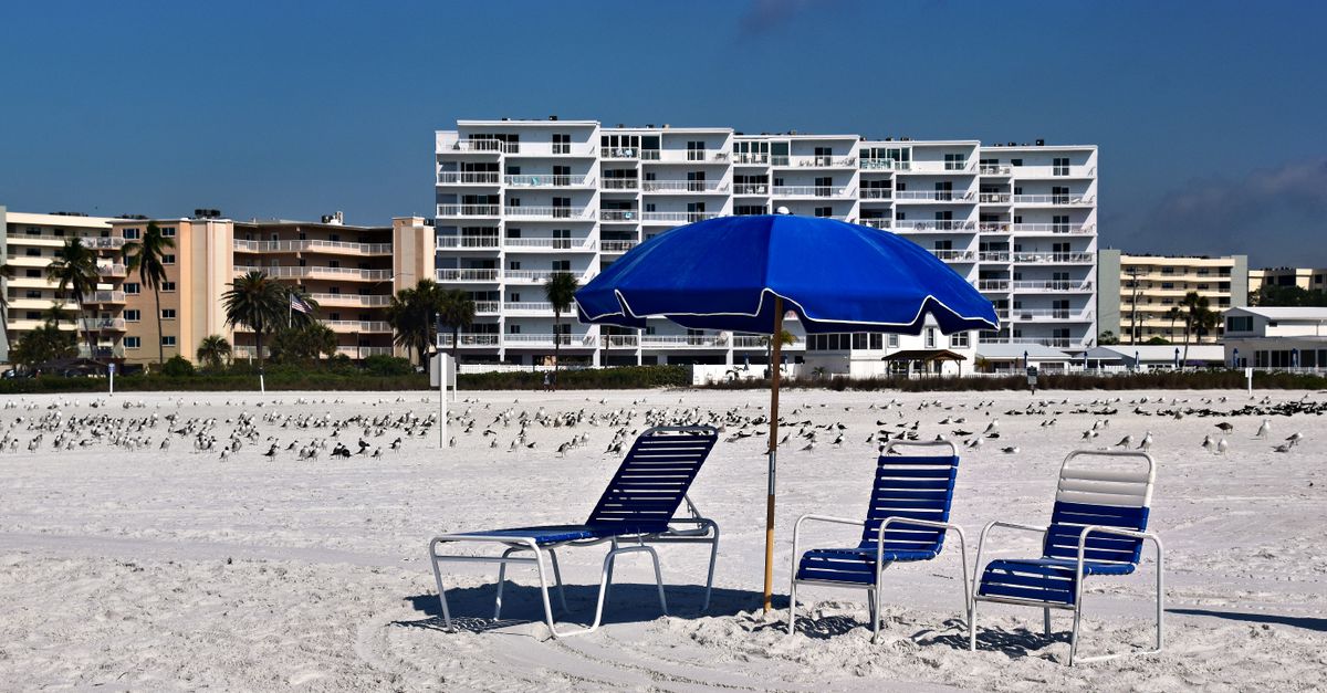 Serene beach scene with a blue umbrella, sun loungers, and buildings in the background.