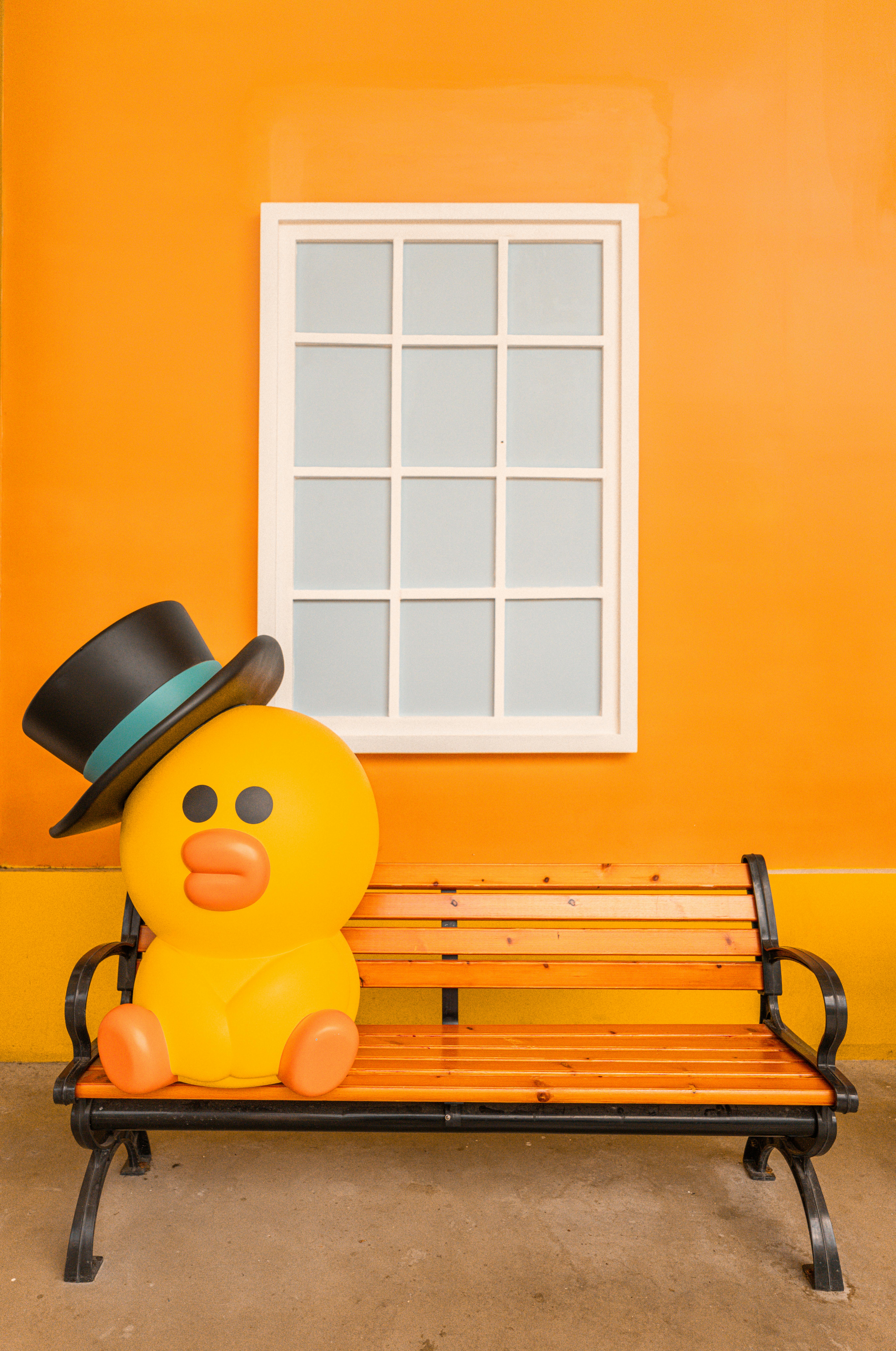 A whimsical yellow rubber duck with a top hat sitting on a bench against an orange wall with a window.