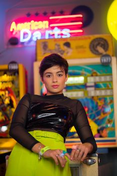 Fashionable portrait of a woman posing in a vibrant, neon-lit arcade with pinball machines.