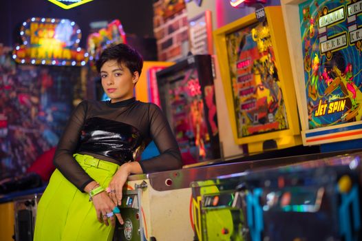 Stylish woman with short hair leaning on pinball machine in vibrant retro arcade setting.