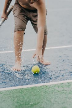 Child interacting with a tennis ball on a rain-soaked court, capturing a playful moment.