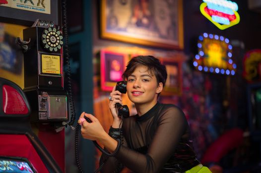 A young woman smiling and posing with a vintage rotary phone in a colorful retro arcade setting.