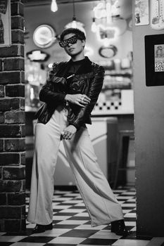 Fashionable woman posing in a retro diner, wearing sunglasses and a leather jacket.