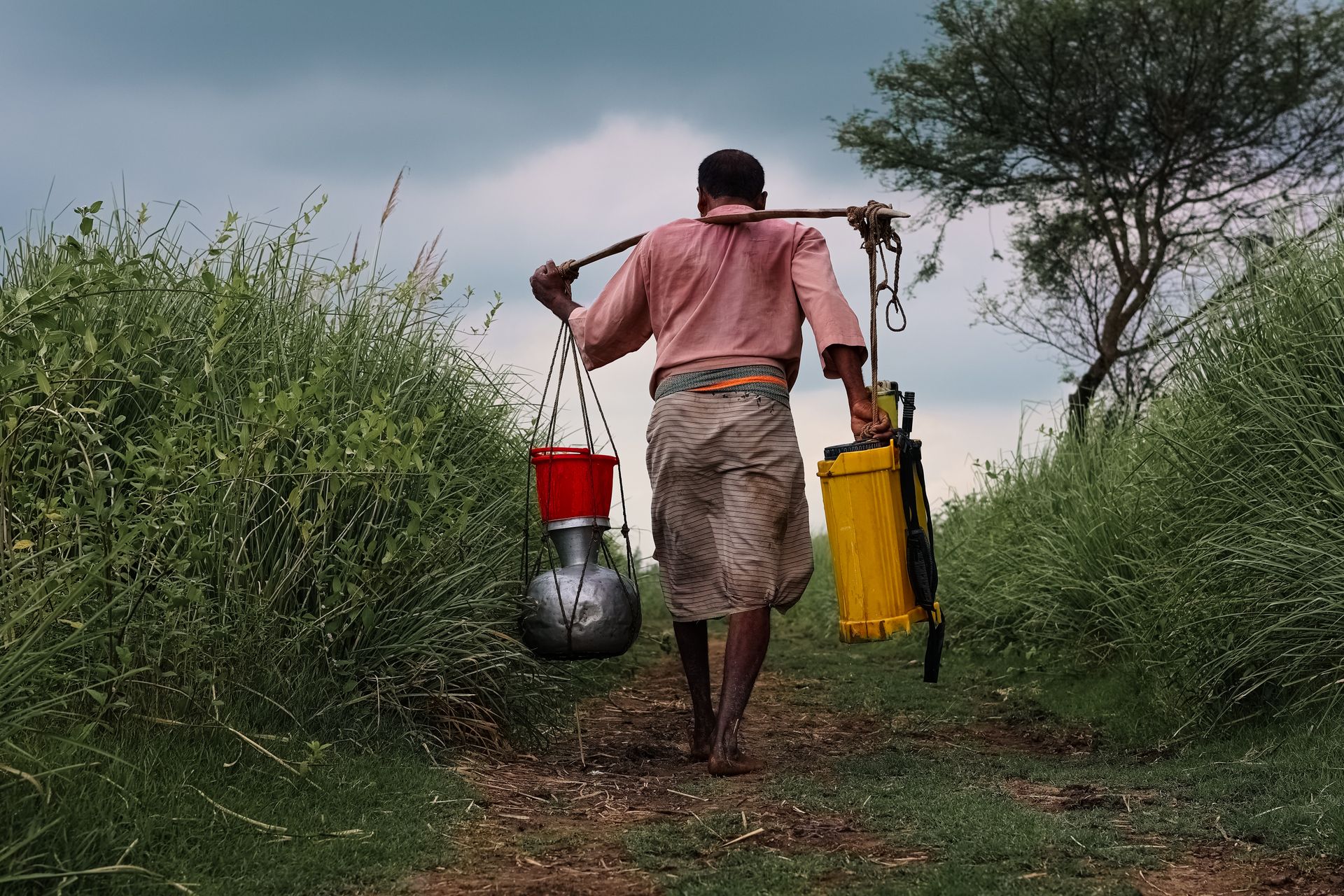 Man carrying tools and buckets on a rural path in Pabna, Bangladesh.