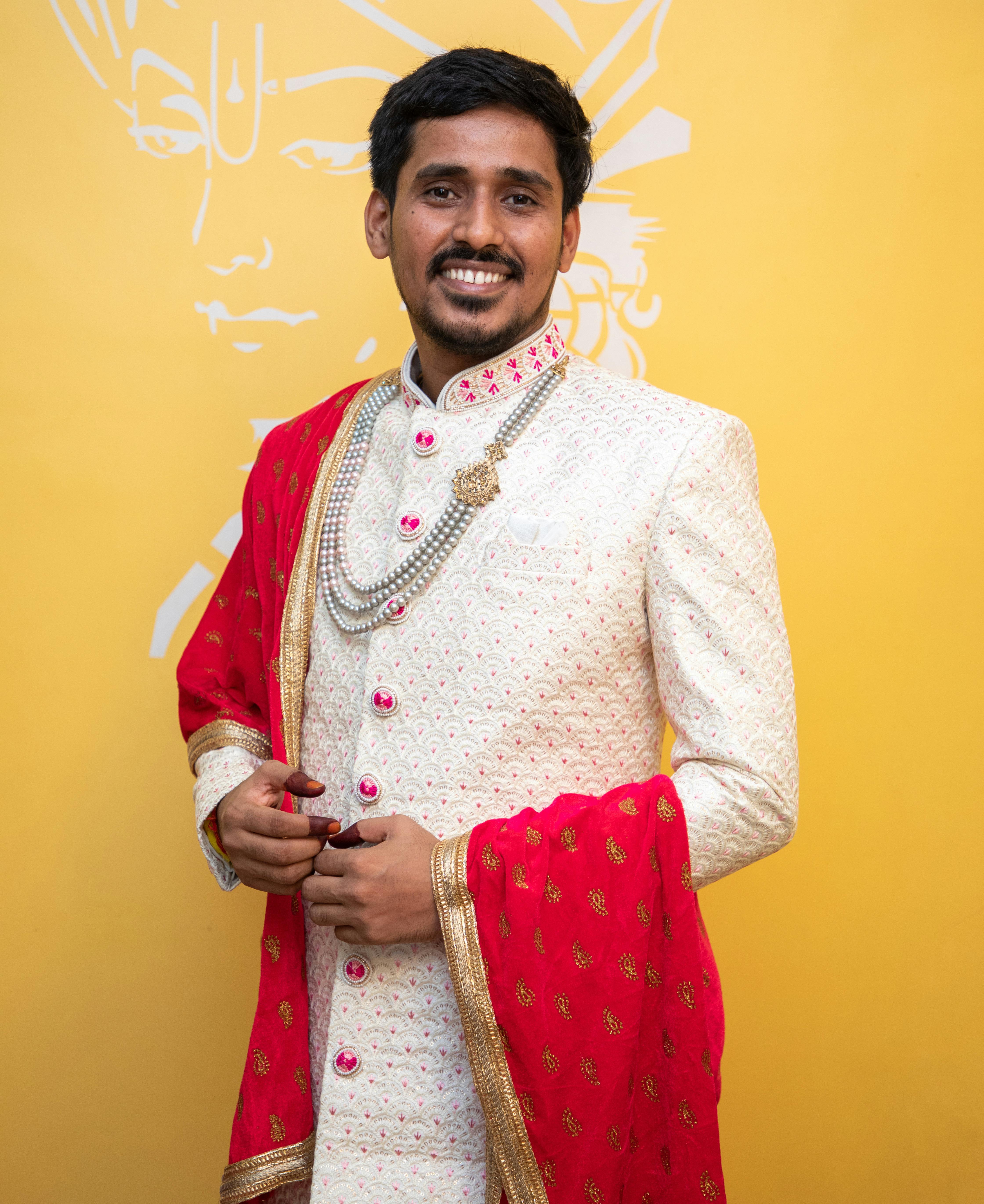 A smiling Indian groom in traditional attire with ornate decorations posing against a yellow background.